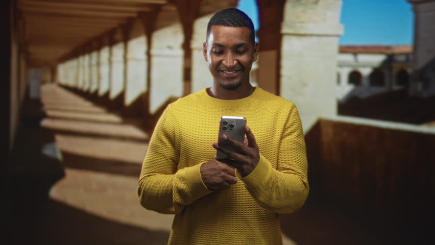 Man in yellow sweater holding smartphone and smiling with hand near mouth in building corridor; joy and connection.