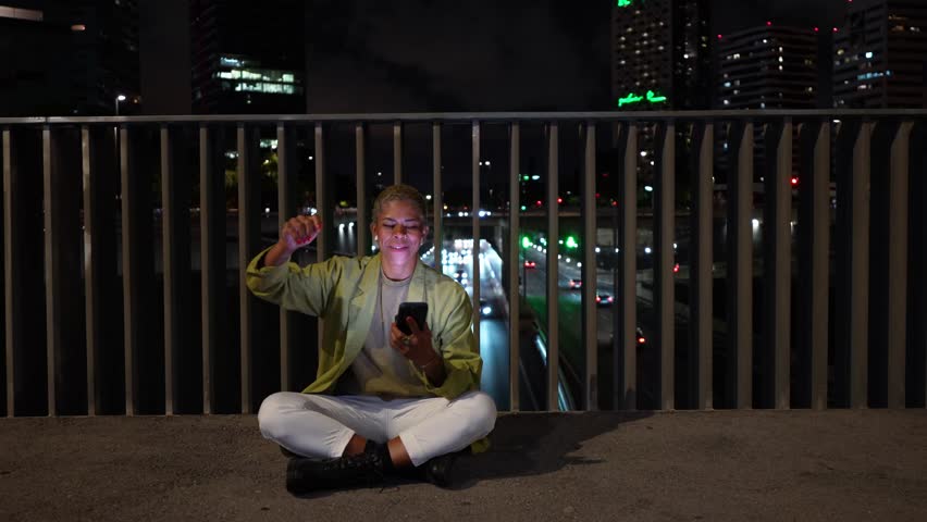 Excited young person with short blond hair sitting on a city bridge at night, celebrating while looking at a smartphone with a raised fist