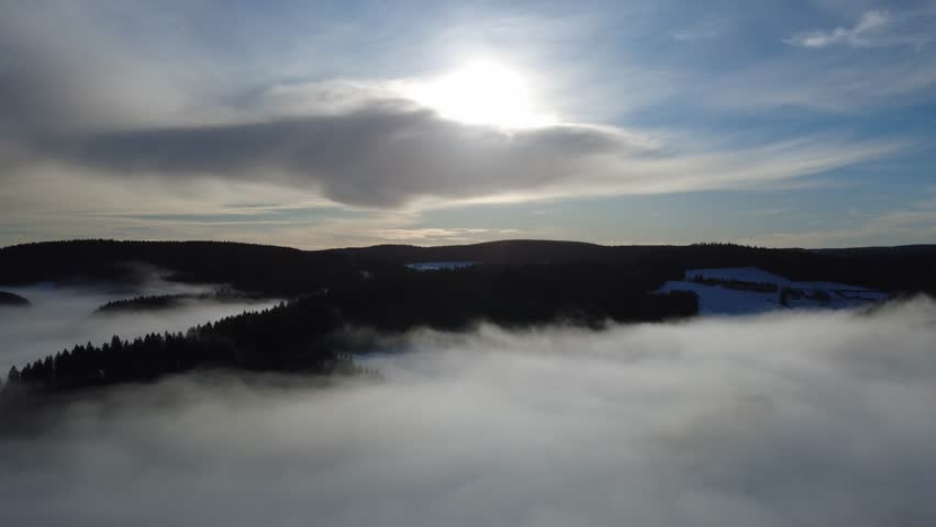 A cinematic drone aerial view of winter fog inversion over the Black Forest hills at sunrise. Misty clouds cover valleys while clear horizon and mountain silhouettes create dramatic atmospheric landscape scenery.