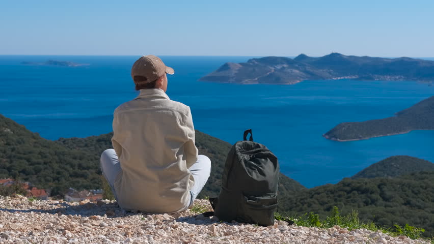 Female hiker enjoying scenic sea view from mountain top and raising her arms.Young woman with a backpack sitting on top of a mountain, raising her arms in a gesture of freedom while admiring the sea