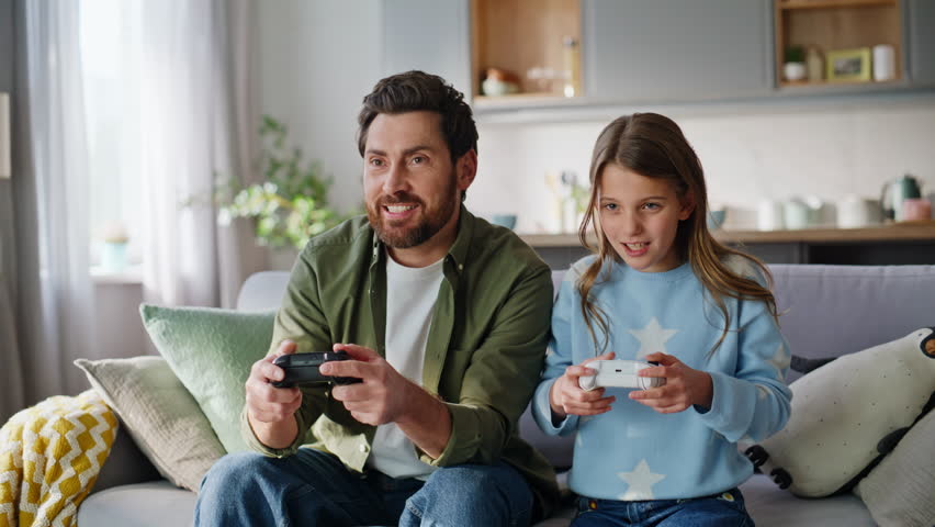 Happy daughter rejoicing online win playing video game with upset father apartment living room closeup. Excited competitive teen girl celebrate victory laugh from dad loser sitting couch together.