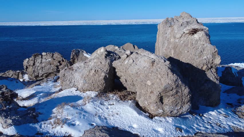 Drone video moving away from a large eroded rock buried in snow and surrounded by sea ice in a polar coastal setting that fades into the horizon. St. Lawrence River, Quebec, Canada.