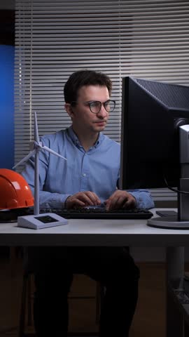 Vertical video male engineer working on a computer in an office, focusing on sustainable energy solutions with a wind turbine model and a hard hat on the desk