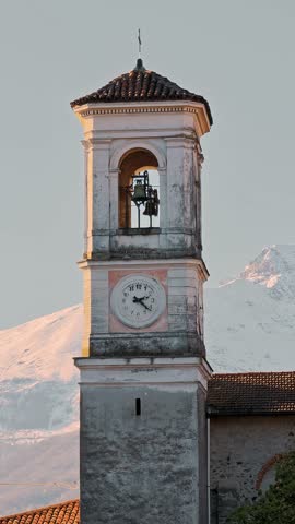 Vertical view of a historic Italian bell tower standing against majestic snow capped mountain peaks. Scenic winter landscape.