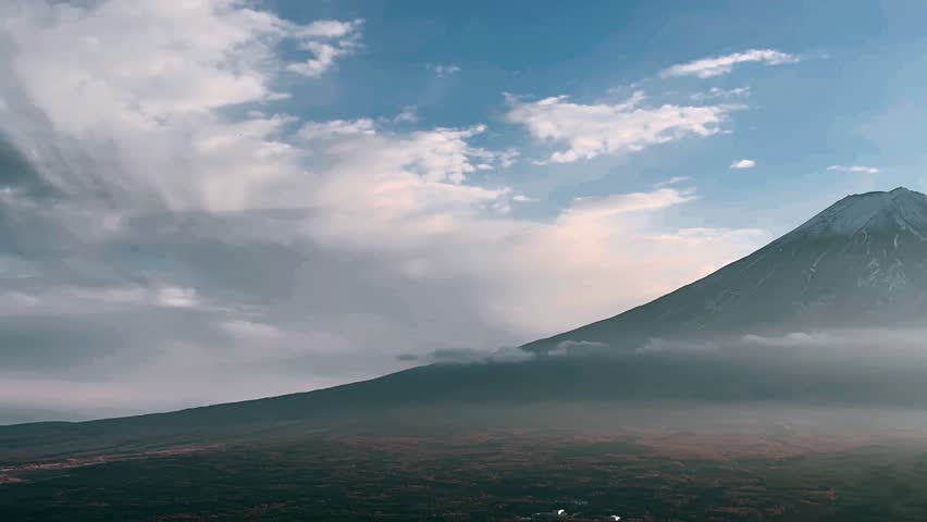 Mount Fuji stands centered in the distance with a light snow cap and faint ridgelines visible down its slopes. A wide band of mist drifts across the lower mountain and the valley below, softening the horizon. Dark pine branches frame the right side of the scene, while scattered clouds sit in a calm pale blue sky.
