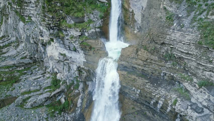 A powerful waterfall plunges down rugged canyon walls, carving through layered rock formations into a clear turquoise pool below. The rushing water creates mist and movement, showcasing the raw beauty of nature and the dramatic landscape shaped over centuries by erosion.