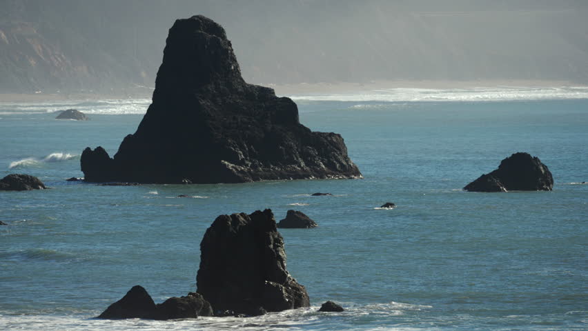 Rocky sea stacks rise from the Pacific Ocean near sandy shoreline, creating dramatic coastal scenery with calm waves, natural formations and open horizon under clear daylight. Oregon