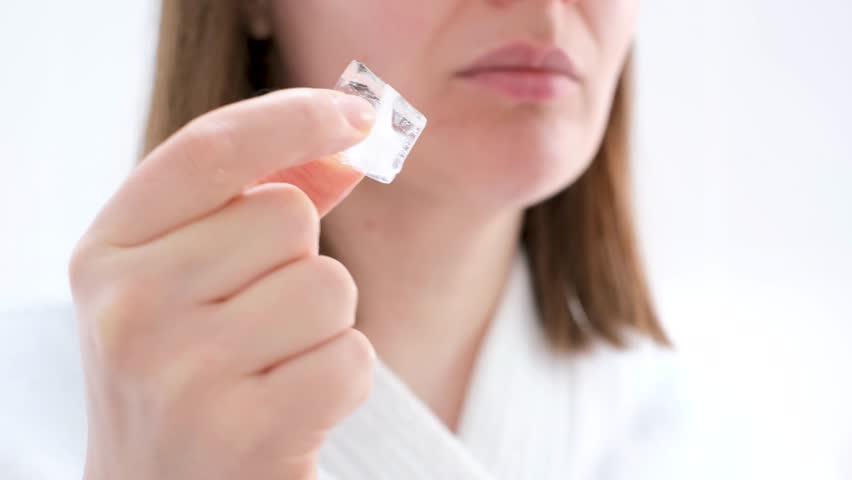 Young woman holding ice cube near her face on white background.