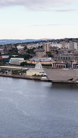 Drone footage captures a contemporary geometric building on a riverfront with a dense urban background.
