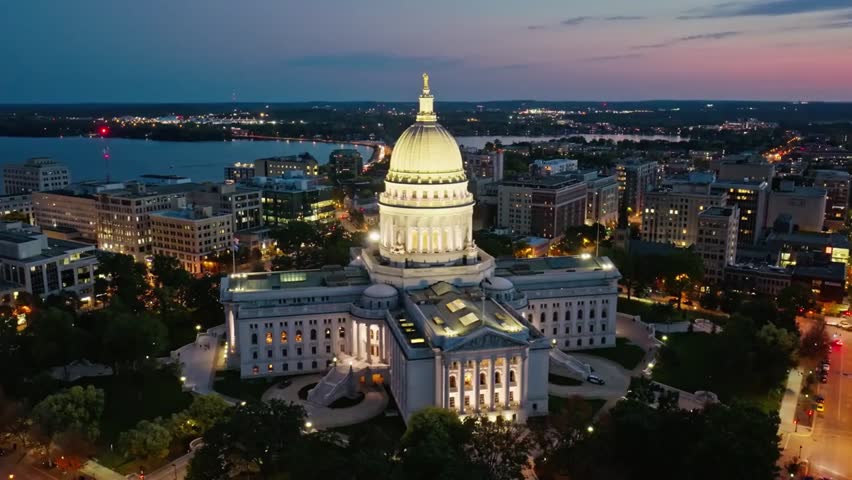 Aerial view of the Wisconsin State Capitol at dusk in Madison, glowing against the evening sky with city lights illuminating the historic skyline.