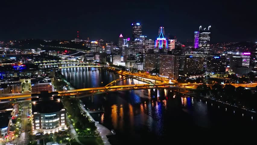 Nighttime aerial view of Pittsburgh skyline with illuminated bridges spanning rivers, city lights reflecting on the water.