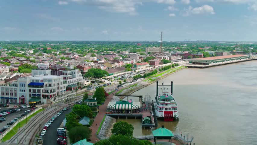 Aerial view of a classic riverboat docked along the Mississippi River in New Orleans, with city skyline and waterfront under clear daylight.
