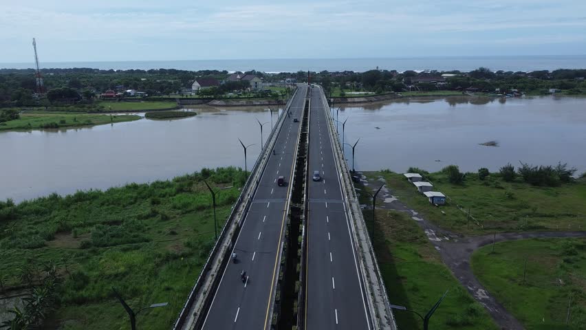 Aerial drone shot flying forward over a coastal bridge near Parangtritis Beach, South Yogyakarta, Indonesia. Scenic view of the quiet beach, ocean, and road with minimal traffic on a sunny day.