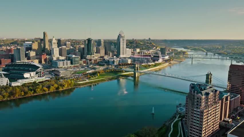 Aerial view of the Cincinnati, Ohio skyline along the Ohio River at golden hour, with city lights and reflections creating a warm, picturesque urban scene.