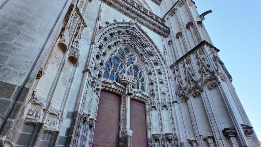 Slow forward movement on Nantes Cathedral entrance followed by gentle tilt up, revealing gothic arches, sculpted columns, ornate stonework and blue sky above.