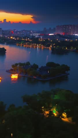 An enchanting aerial panorama of Daming Lake at dusk, showcasing vibrant sunset hues over the tranquil water. Illuminated traditional architecture on islands and city skyline lights create a mesmerizing and serene evening landscape.