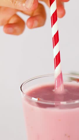 Close-up vertical shot of a hand stirring thick pink smoothie with a red-and-white straw.