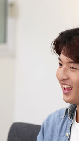 A cheerful young Asian man in a denim shirt talks and points upward indoors