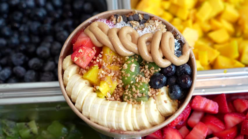 Top down view of a delicious and healthy acai smoothie bowl being prepared with fresh banana, strawberry, blueberry, kiwi, and mango, topped with peanut butter, granola, and coconut flakes