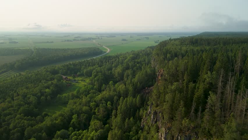 Drone aerial flyover glides along dramatic Hunneberg cliff edge revealing dense pine forest canopy winding road through green farmland and panoramic misty views on serene summer day in Sweden