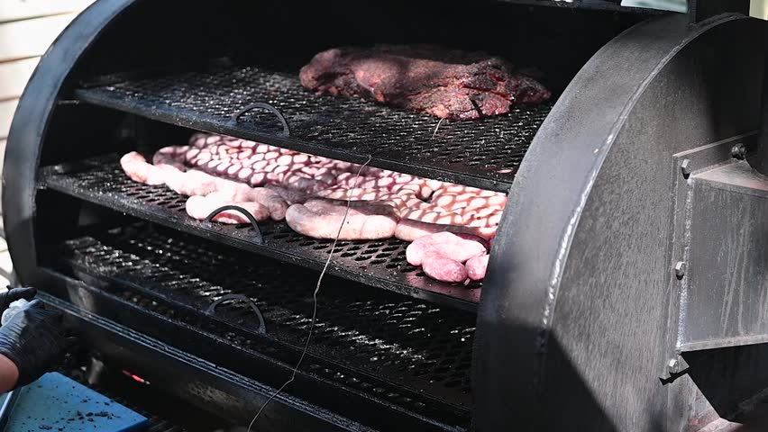 A grill is set up outdoors with multiple racks cooking various cuts of beef and sausage. The smoke rises from the grill, indicating the cooking process.