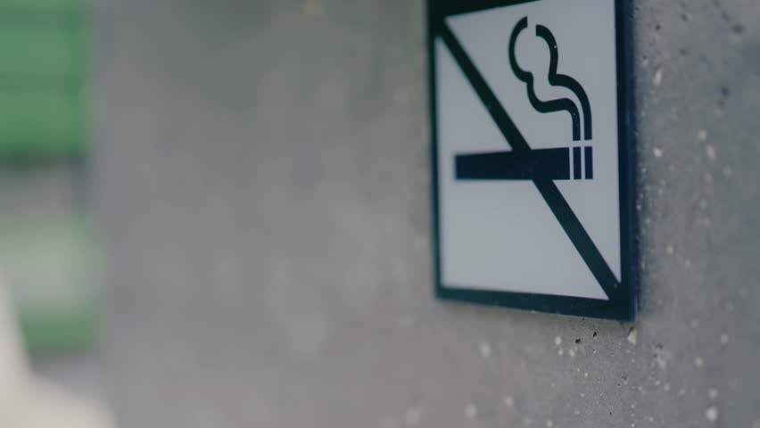 Close-up of a no smoking sign sticker on a public concrete wall. Indicating a smoke-free area with a blurred background showing a park or outdoor space. A symbol of prohibition and healthy lifestyle