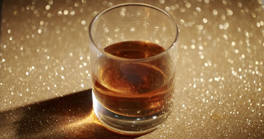 Golden whiskey swirling with ice cubes in a rocks glass, filmed in slow motion and isolated on a golden background. Golden whiskey glass with ice cubes stirring on a sparkling background