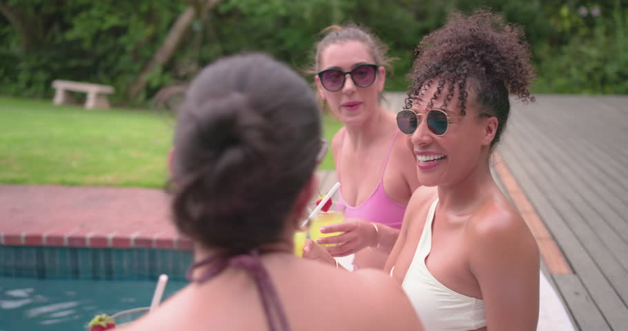 Diverse female friends in bikinis smiling at camera clinking cocktails relaxing poolside splashing. Pool, backyard, swimwear, sunglasses, deck, summer, candid
