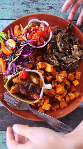 Colorful plate of plant based food including sweet potatoes, mushrooms, and fresh salad being placed on a rustic wooden table by a person, representing a healthy vegetarian lifestyle