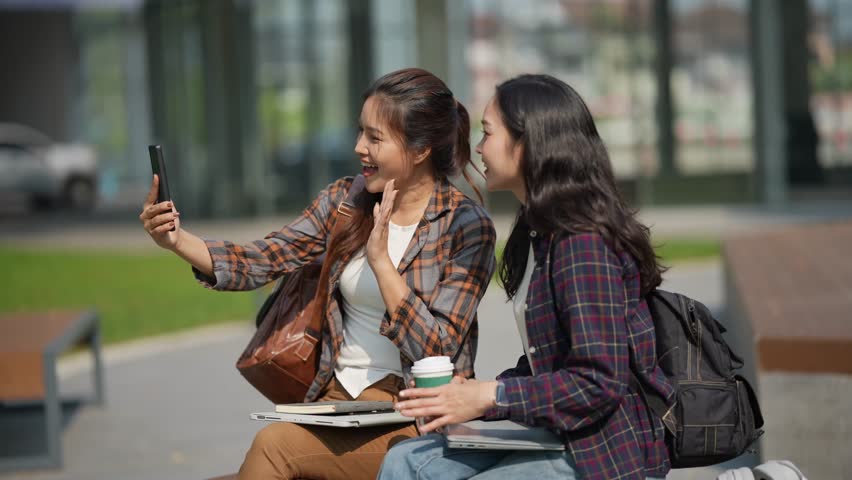 Two smiling Asian female students sit on a bench in a university park with their laptops, studying, technology, and people.