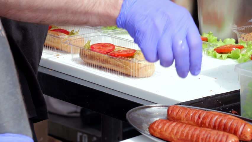 Chef prepares sausage, Cook assembles sausage in bun, Food vendor arranges sausage in fresh bread, Caterer carefully places hot sausage inside bread roll with garnishes