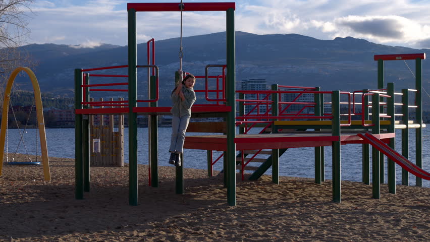 Young girl in cold weather clothing riding on a zipline located at a lakeside playground, coming towards the camera with a big smile.
