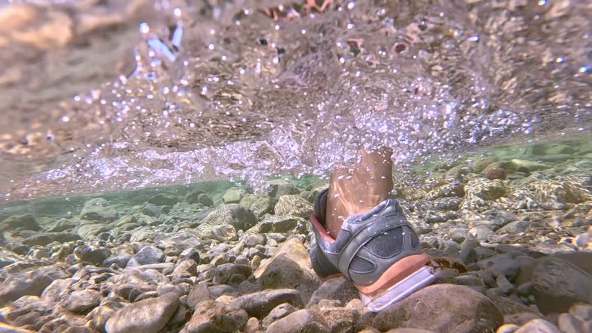 CLOSE UP, UNDERWATER, LOW ANGLE: Female person stepping through a shallow rocky river, sending water droplets into the air. Clear stream, pebbled riverbed, green forest and summer outdoor adventure.