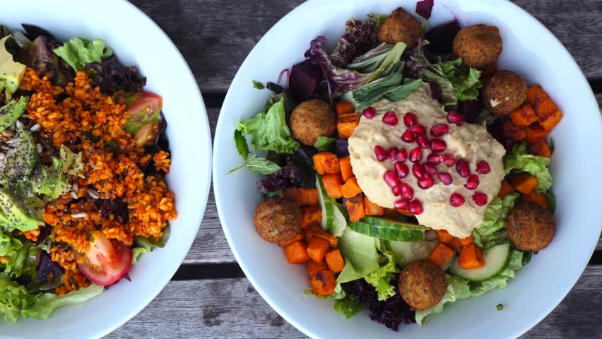 Two vibrant and delicious vegan salads sitting on a rustic wooden table, one featuring falafel, hummus, and pomegranate seeds and the other with avocado, quinoa, and tomatoes for a healthy meal