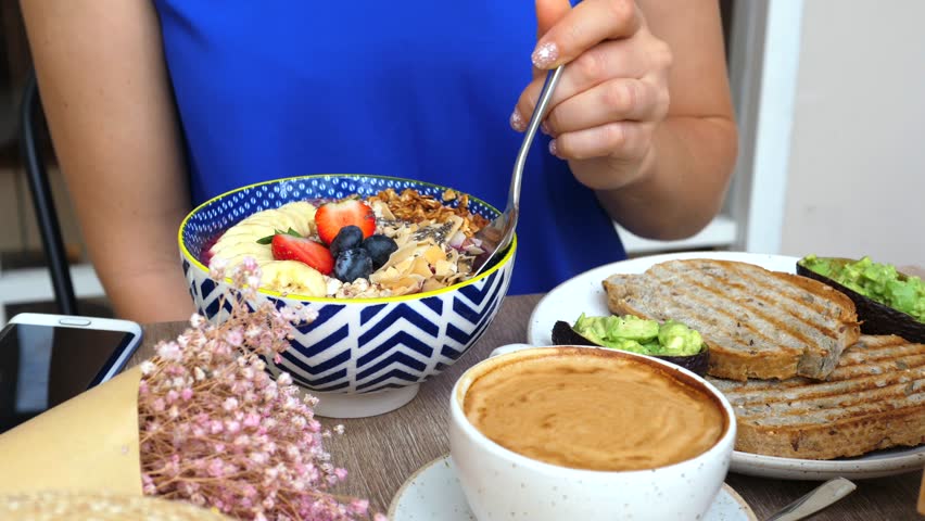 Unrecognizable woman in a blue top eating a healthy smoothie bowl with fresh fruits, berries, and granola for breakfast in a cafe, along with avocado toast and a cup of coffee