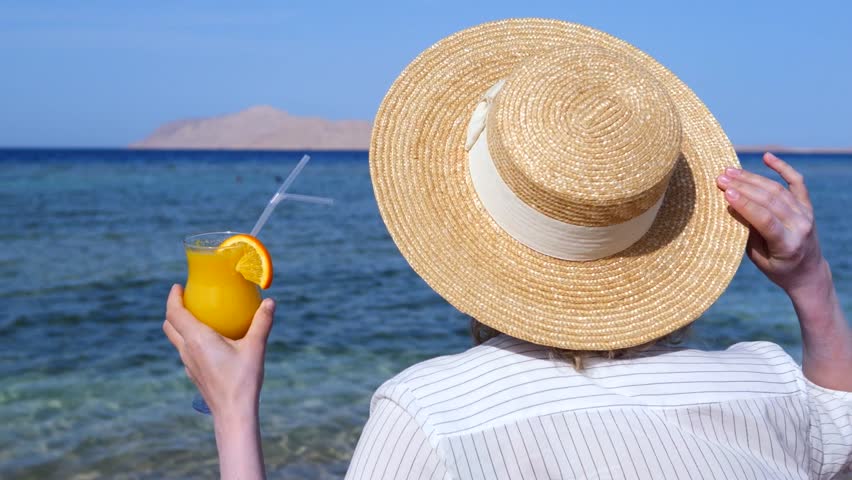 Back view of a young woman wearing a straw hat and holding a glass of fresh orange juice with an orange slice, enjoying the beautiful sea view on a sunny day during her summer vacation