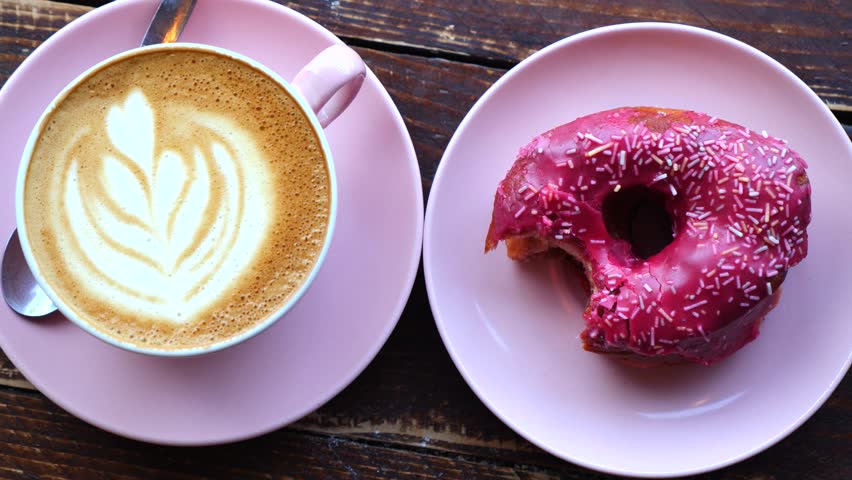 Delicious breakfast scene featuring a pink cappuccino cup with latte art and a matching plate with a pink glazed donut being eaten, all set on a rustic wooden table in a cafe