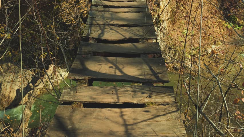 A high-angle, first-person view of a long wooden suspension bridge. The rustic path leads through a dense autumn forest, evoking a sense of adventure, exploration, and tranquility
