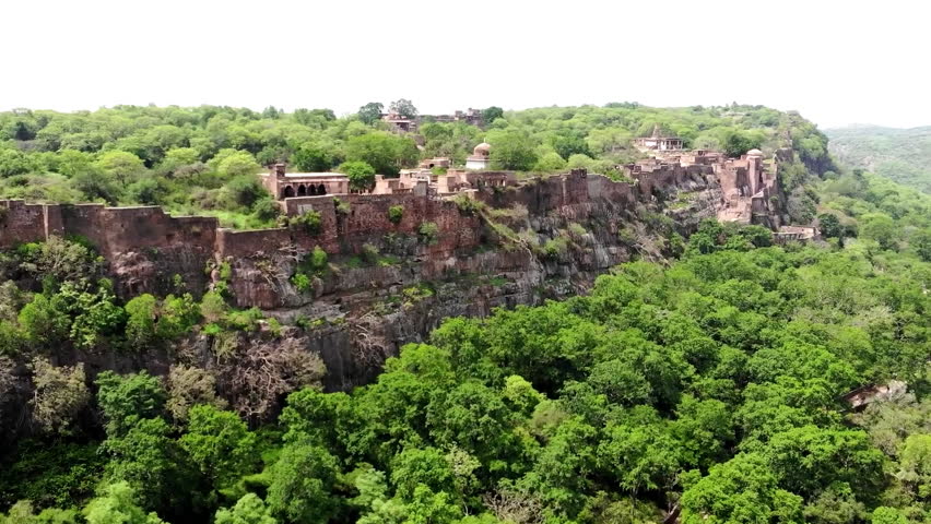 A cinematic aerial shot overlooking the historic Ranthambore Fort, a UNESCO World Heritage site situated within the Ranthambore National Park in Rajasthan, India. The footage captures the sprawling sandstone ramparts and ancient temples perched atop a high plateau, surrounded by the rugged forest canopy and lakes that define one of India