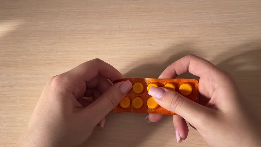 Pressing medicine tablet from blister pack and dropping pill onto wooden table closeup pharmaceutical dosage healthcare treatment concept pharmacy medication routine