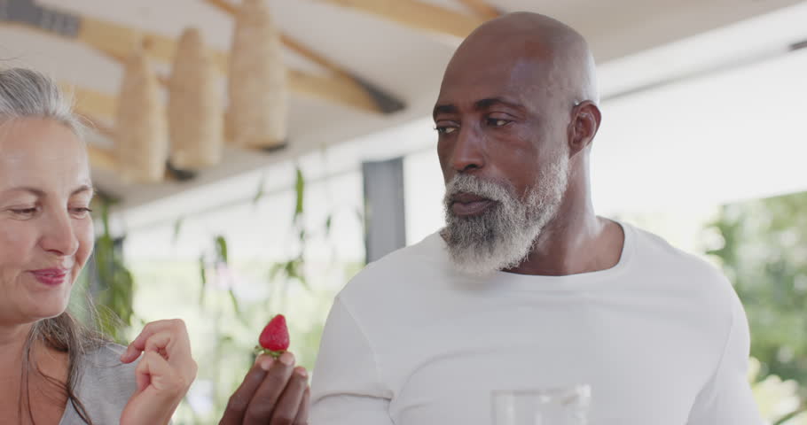 Senior couple on porch man offering strawberry sparking affection, woman leaning in, kissing finger. Outdoor, veranda, daylight, greenery, lanterns, wicker, beams