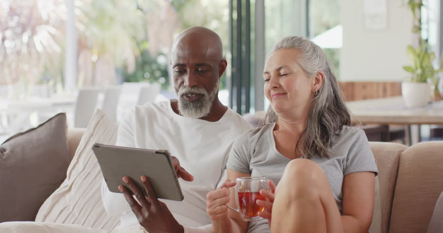 Diverse couple on sofa, senior man tapping tablet, sharing photos with partner with mug. Livingroom, cozy, screen, home, sunlit, plants, patio