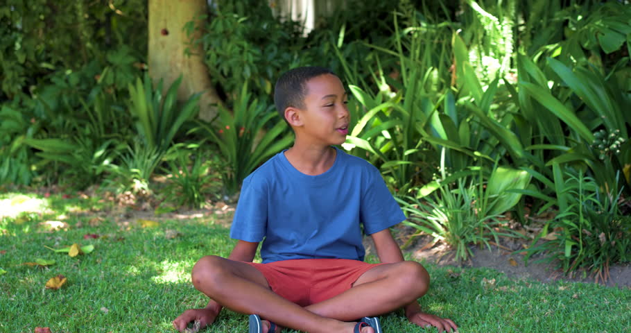 African American child sitting on lawn in blue tee board nearby noticing something above smiling. Youth, outdoor, backyard, tree, plants, skateboard, sunshine