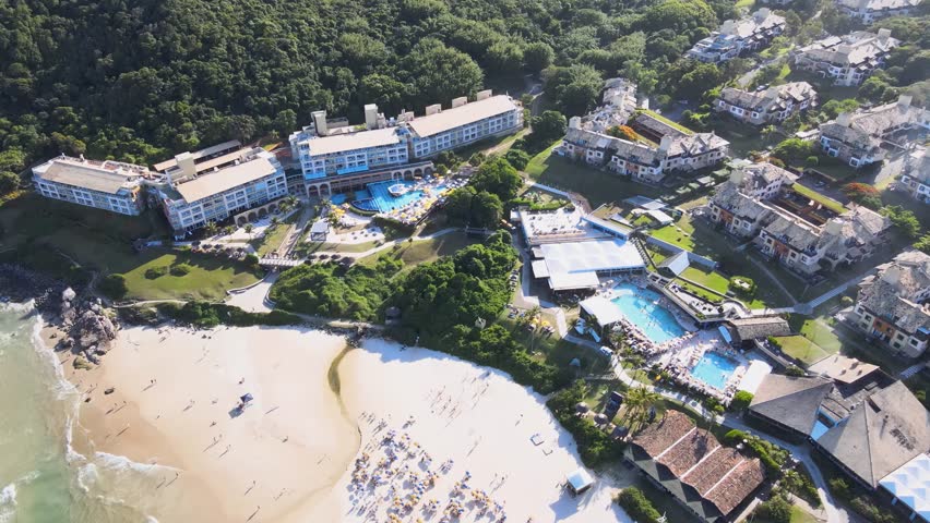 High angle aerial view of the luxury Costao do Santinho Resort in Florianopolis, Brazil, showing hotel buildings, swimming pools and crowded beach on a sunny summer day.
