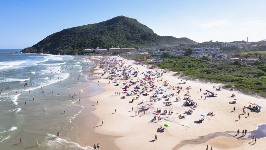 Low-altitude aerial wide shot of a crowded Santinho Beach in Florianopolis, Brazil, featuring tourists, colorful umbrellas and the green Morro das Aranhas mountain on a sunny day.
