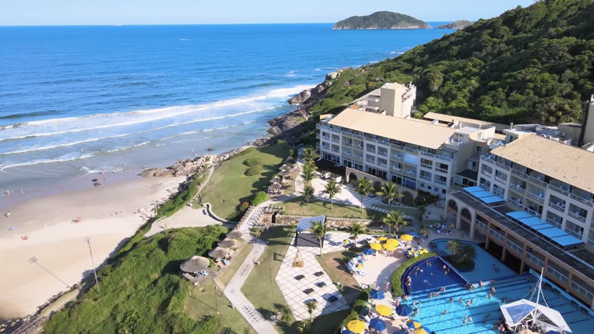 High angle aerial view of Costao do Santinho Resort in Florianopolis, Brazil, showcasing hotel buildings, swimming pools and rocky cliffs by the Atlantic Ocean on a sunny day.
