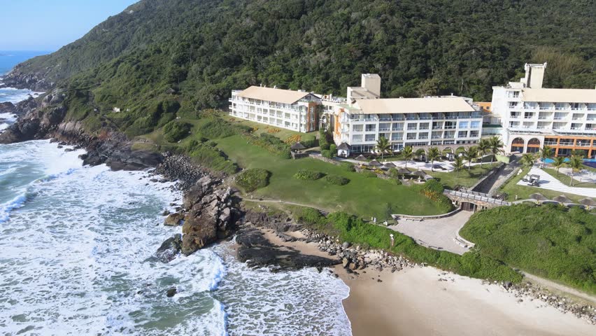Cinematic aerial view of the luxury Costao do Santinho Resort in Florianopolis, Brazil, showing hotel buildings by the ocean waves and green mountains on a sunny day.
