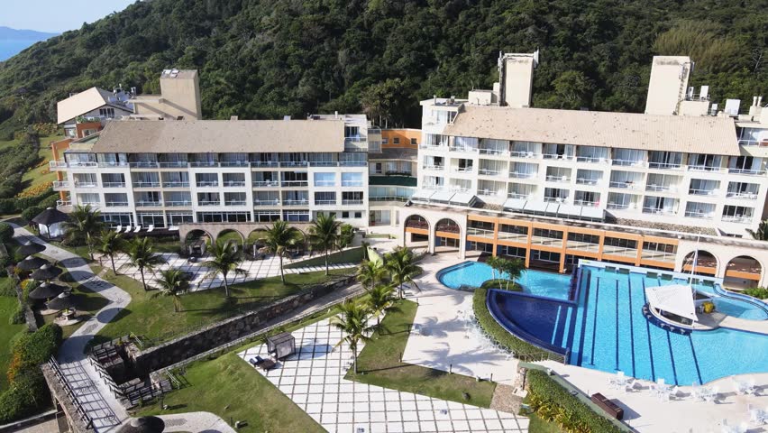 Cinematic high angle aerial view of luxury swimming pools and hotel buildings at Costao do Santinho Resort in Florianopolis, Brazil, with ocean waves and green mountains on a sunny day.
