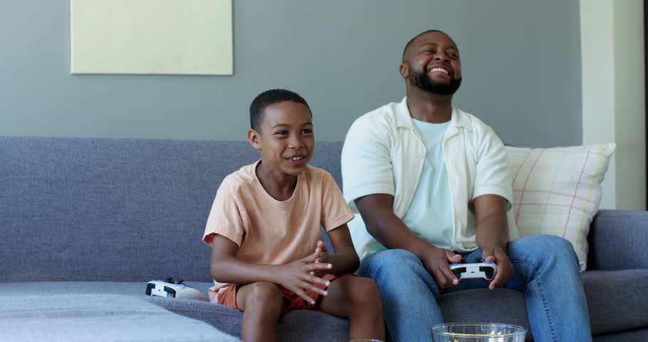 African American father and teen son gaming on couch, son standing, celebrating controller win. Family, playtime, celebration, laughter, sofa, home, casual