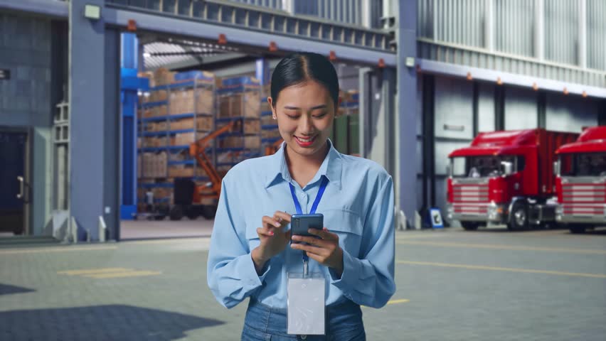 Asian Female Professional Worker Standing With Her Smarphone, Outside of Logistics Distributions Warehouse, Work Continuously On Her Device